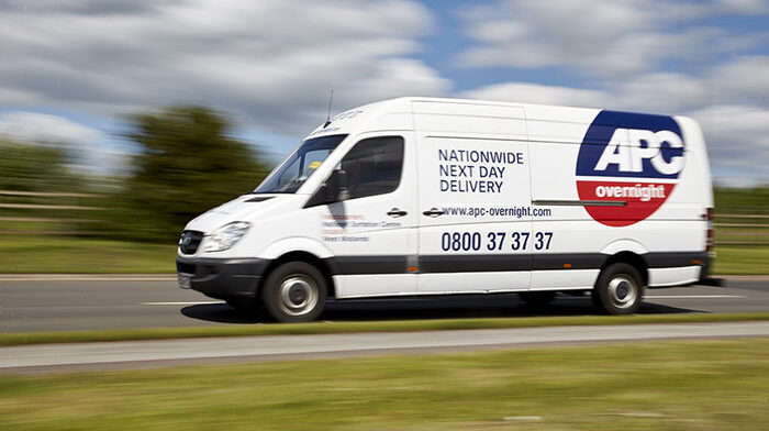 An APC network delivery van driving down a rural road with green grass in front and a cloudy blue sky behind.