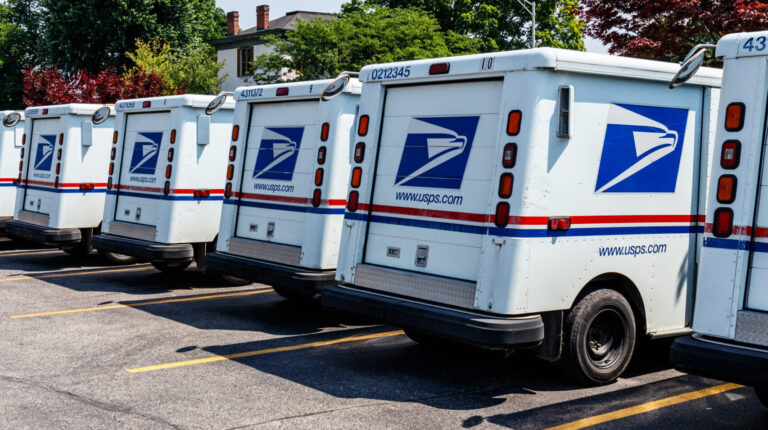 A row of USPS mail vans parked, with green trees in the background