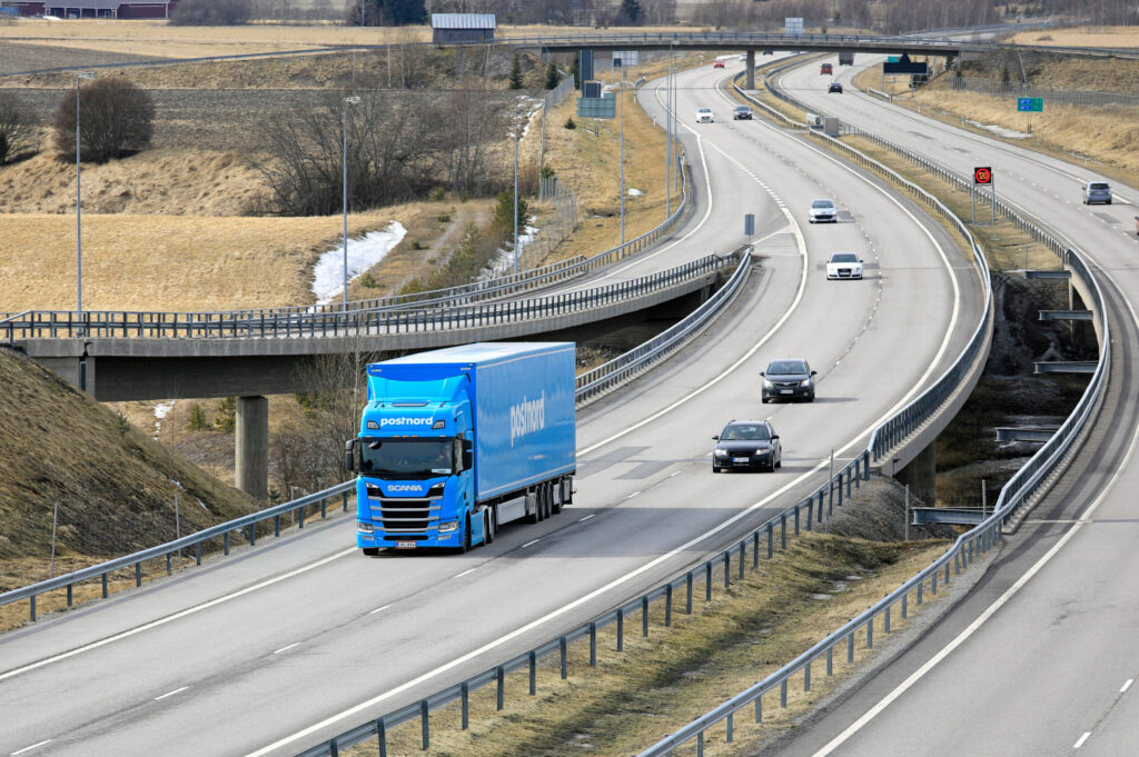 Springtime landscape of European Route E18 in South of Finland with Next Generation Scania R500 semi trailer of FE-Trans for PostNord Logistics among motorway traffic.