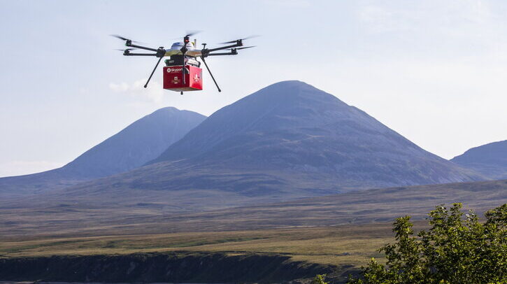 Drone with red Royal Mail parcel flying over lake with mountains in background