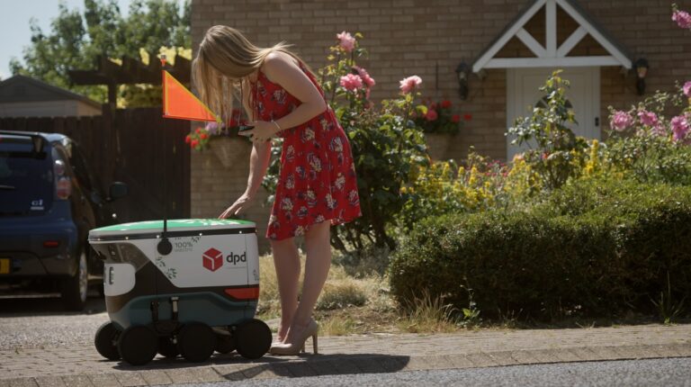 Woman in red dress outside residential house collecting item from DPD autonomous robot using mobile phone