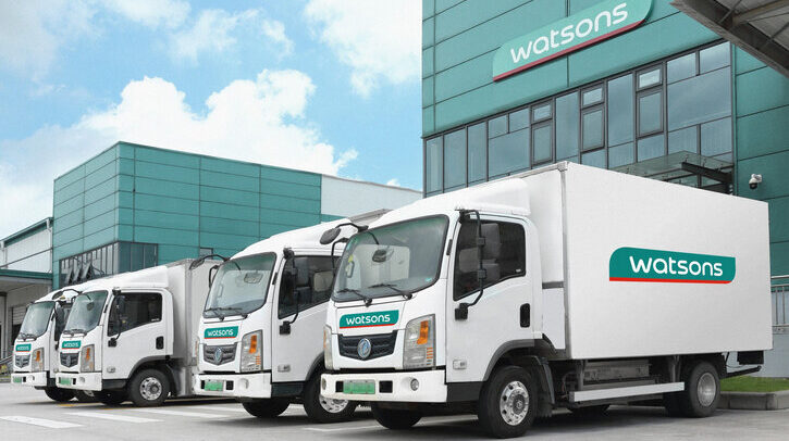 White delivery trucks with green Watsons logo on the side, in front of building with blue sky and white clouds in background