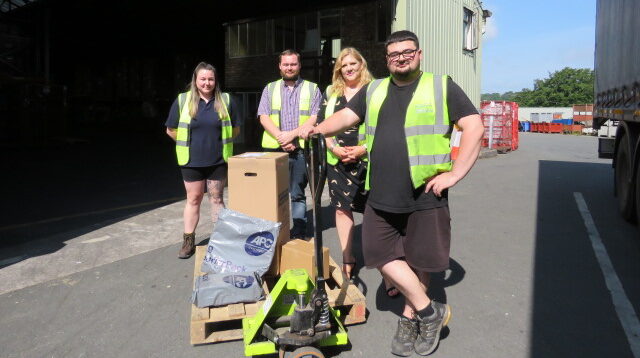 Four logistics workers in high-vis jackets stand in front of a pallet and warehouse.