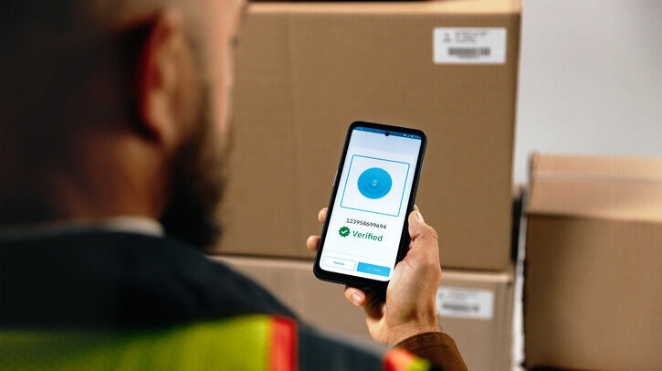 Logistics worker wearing a hig-vis jacket uses a handheld mobile device in a warehouse of brown cardboard boxes.