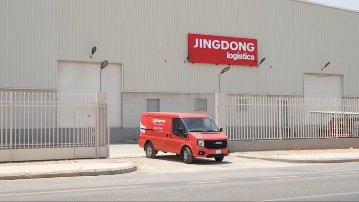 JD Logistics van in front of a grey JD warehouse. The vehicle is exiting the building and turning onto a empty road.