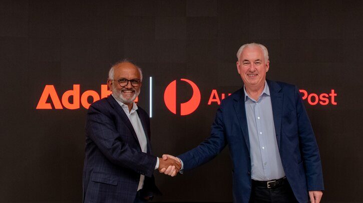 Shantanu Narayen, Adobe chair and chief executive officer, and Paul Graham, CEO and managing director, Australia Post, shaking hands in front of a background with the company logos on, marking their new partnership