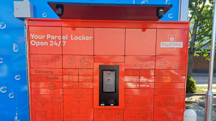 A bank of exterior red parcel lockers with the Royal Mail logo on the top right and a blue wall behind