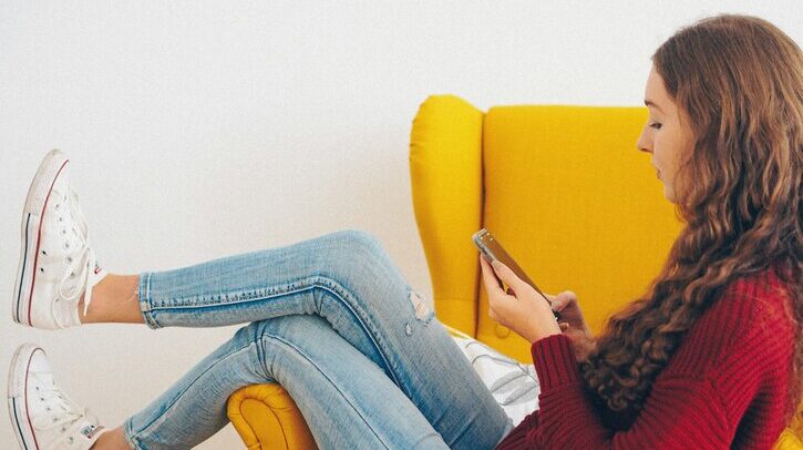 A woman in a red jumper and blue jeans reclines on a yellow sofa and looks at her smartphone while make an e-commerce order.