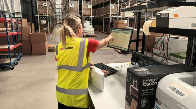 An XPO Logistics team member in a yellow high-vis jacket handles a package while working on a digital touchscreen in a warehouse