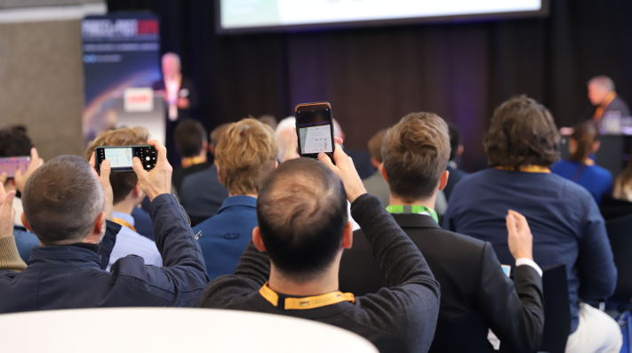 People sit in rows at a workshop at a conference listening to a speaker while taking photos with their cellphones