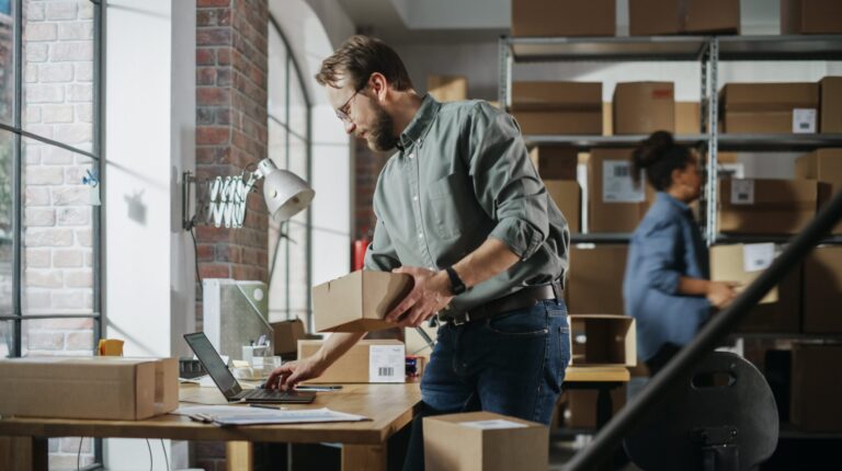 A man prepares a small parcel for sending. He is standing in an exposed brickwork warehouse using a laptop