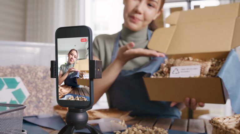 A woman performs an unboxing for the camera in an apartment during the day, with a smartphone on a stand on a table and a window behind her