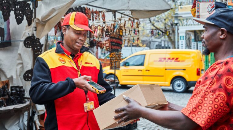A DHL worker scans a parcel in a busy street with a yellow, branded DHL van parked behind