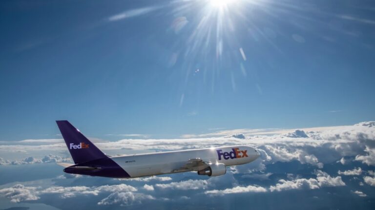 A FedEx cargo aircraft flies above the clouds with the sun shining in the background