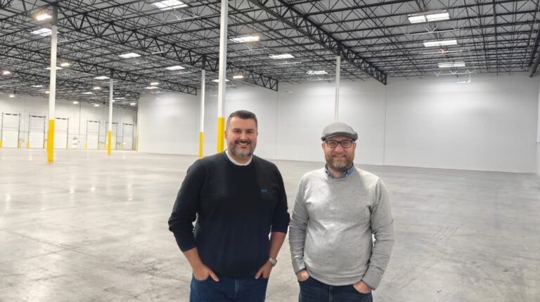 Two men stand next to each other in Fidelity Fulfifment's new Salt Lake City warehouse facility. The floor space is empty and the ceiling high, with strips of lighting and columns with yellow high vis markings around their lower half.