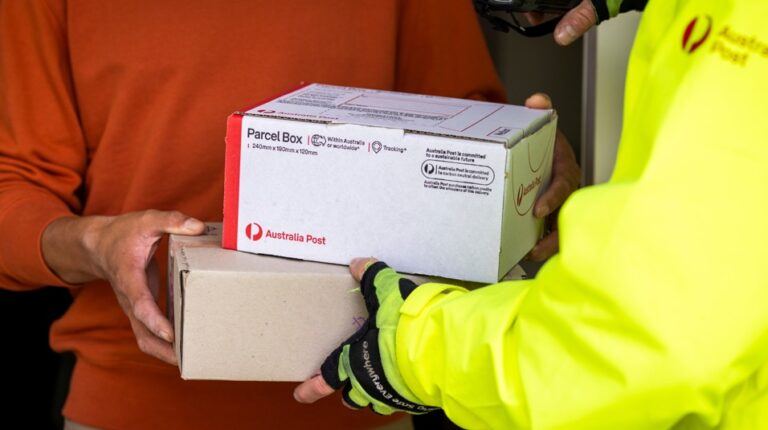 An Australia Post worker wearing a yellow high-vis jacket delivers a parcel to a customer, who is wearing an orange-red jumper. The photo shows their torsos but not their faces.
